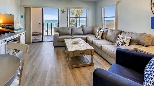 Ein Wohnzimmer mit Sofa, Couchtisch und Fernseher, ein Balkon mit Blick auf den Strand und ein Essbereich mit Tisch und Stühlen.