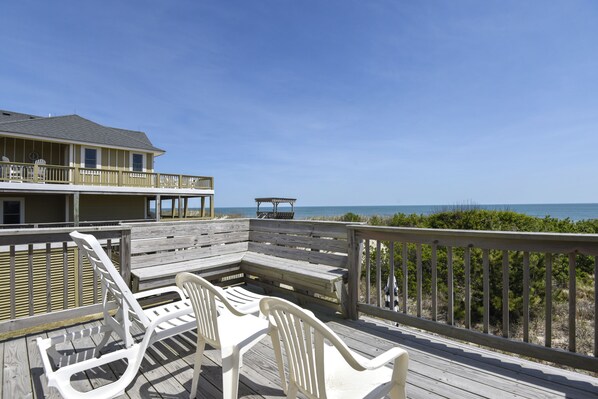 A wooden deck with white plastic chairs and a bench, overlooking a beach and a building with a balcony. - WH1019- Oceanfront Oasis! Featuring 3 BRs, Wifi, Ocean Views and Hot Tub!! (Corolla)