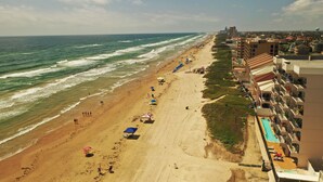 A beach with a long stretch of sand, a pool area, and buildings along the shore.