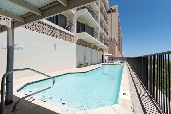 A swimming pool with a metal ladder, a covered area with a white ceiling, and a building with balconies in the background. - Florence II 403 - Rattan resort vibes;Cute bedrooms;Beachfront pool (South Padre Island)