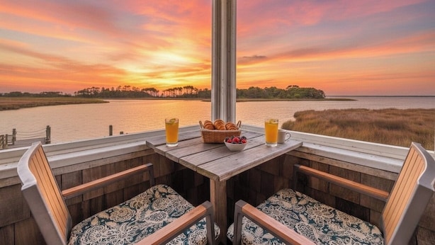 A wooden deck with two chairs and a table set for breakfast, overlooking a body of water and a sunset.
