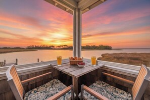 A wooden deck with two chairs and a table set for breakfast, overlooking a body of water and a sunset.