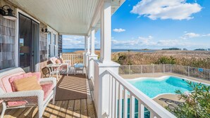 A covered outdoor area with a pool, white railings, wicker furniture, and a view of the beach.