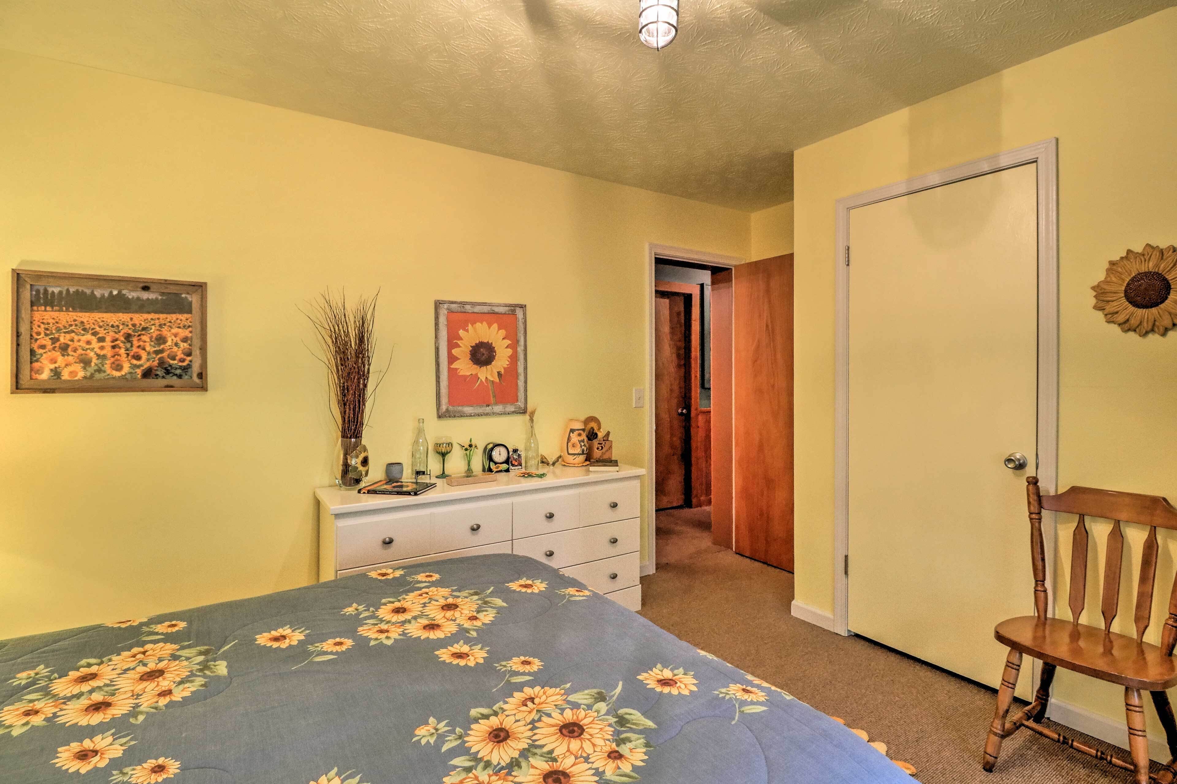Een slaapkamer met een bloemenbedekking, een dressoir met decoratieve objecten, een stoel en een deur die naar een andere kamer leidt.