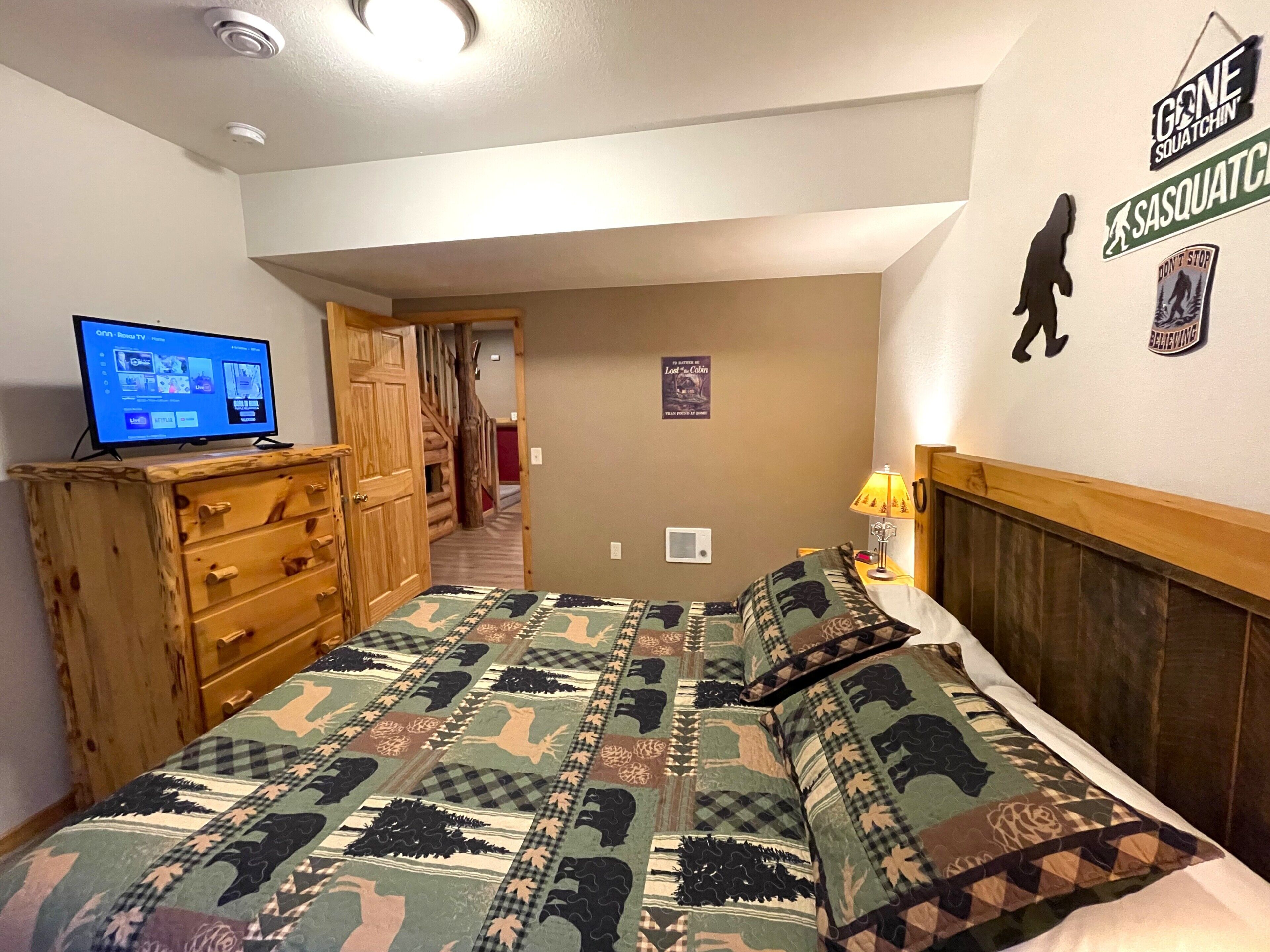 A bedroom with a bed, a TV on a dresser, a wooden dresser, a wall with motivational posters, and a hallway leading to another room.