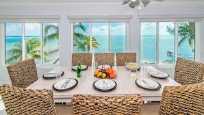 A dining area with a view of the ocean, wicker chairs, a table set for four, and a ceiling fan. - Blue Serenity: Beach House w/Pool, Pickleball Courts + Backyard Snorkeling (Grand Cayman)