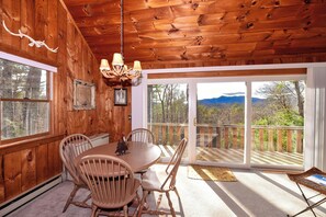 A wooden interior with a dining table and chairs, a chandelier, and large windows with a view of trees and mountains.