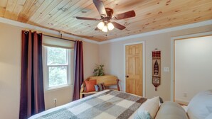Une chambre avec un plafond en bois, un lit recouvert d’une couverture à carreaux, un ventilateur de plafond et une fenêtre avec des rideaux.