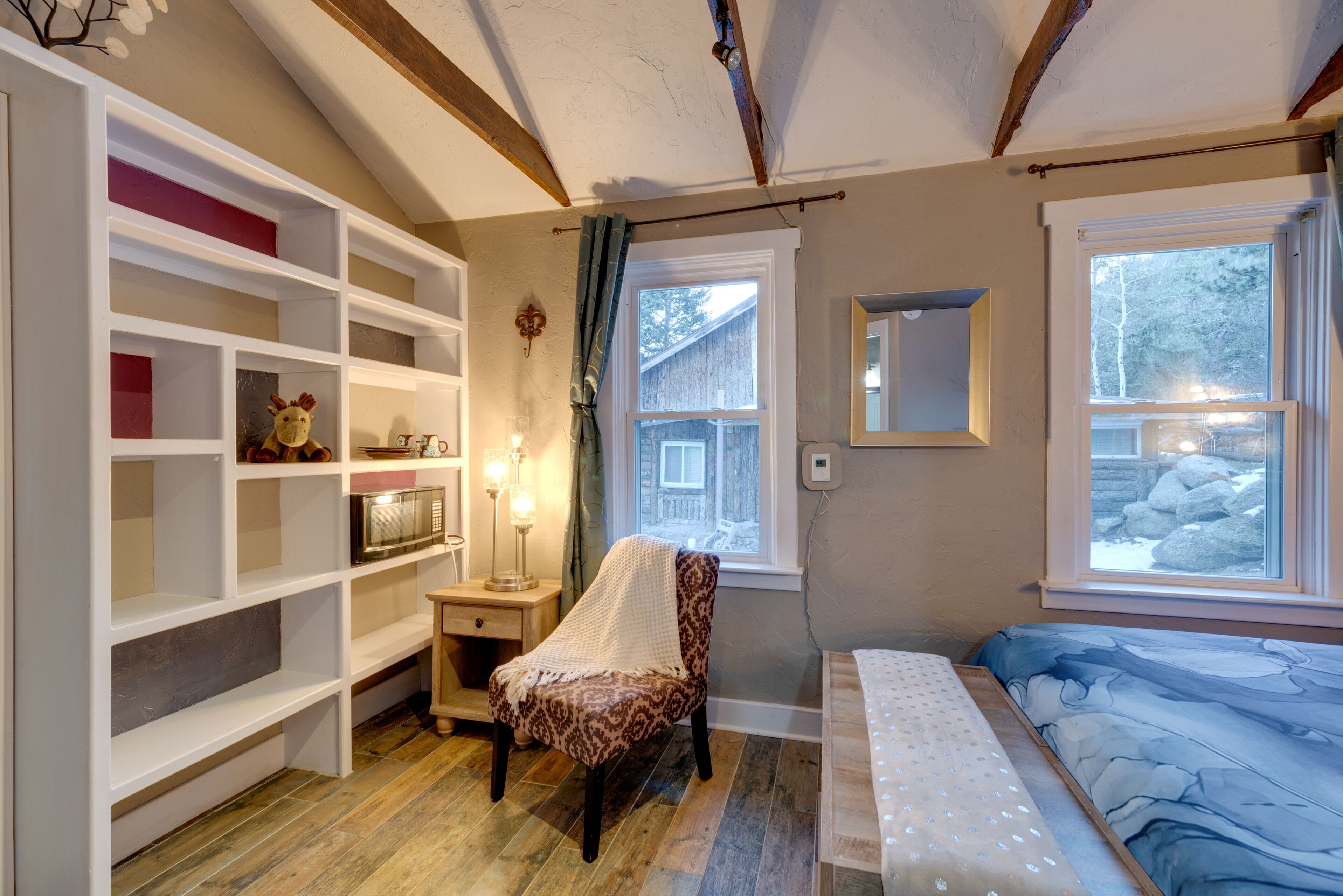 A cozy bedroom with a bed, a chair, a shelf unit, and a window with a view of trees.