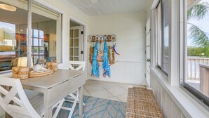 Een overdekt patio met een eettafel, stoelen en een bankje. Op de tafel staan rieten manden, het bankje is geweven en aan de muur hangt een kapstok met daaraan jassen en andere kleding.