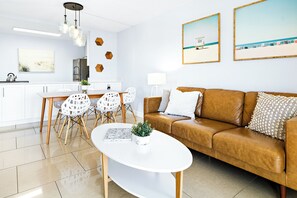 A modern living room with a brown leather sofa, white coffee table, and a dining area with white chairs and a wooden table.