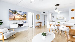 A modern living room with a flat-screen TV, a white coffee table, and a dining area with a wooden table and white chairs. - Pier-Side Penthouse Spacious Corner Unit W/Heated Pool & BBQ Grills! 🌴 (Cocoa Beach)