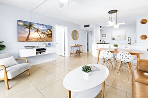 Una sala de estar moderna con un televisor de pantalla plana, una mesa de centro blanca y un comedor con mesa de madera y sillas blancas.