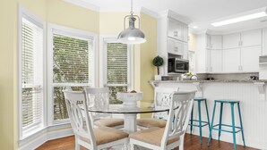A kitchen with white cabinets, a glass-top dining table, and two blue bar stools. - Ocean Mist Cottage | Steps to Beach | Private Pool | Charming Neighborhood (Destin)