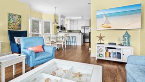 A living room with a blue armchair, a coffee table with seashells, a kitchen in the background, and a beach-themed painting on the wall. - Ocean Mist Cottage | Steps to Beach | Private Pool | Charming Neighborhood (Destin)