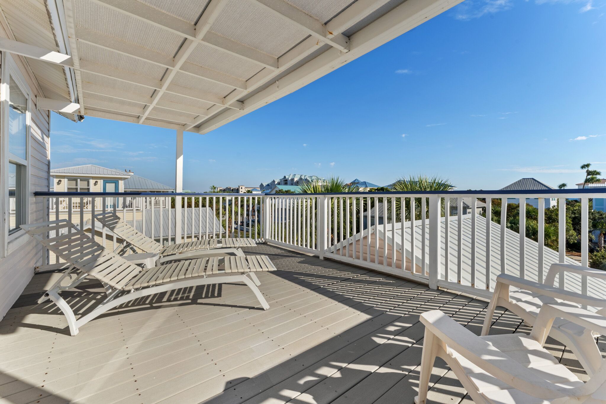 A covered outdoor area with white railings, wooden lounge chairs, and a view of buildings and palm trees.