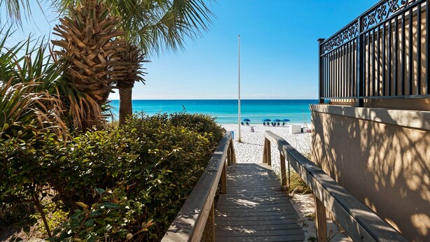 A wooden pathway leading to a beach with a palm tree and a fence on the right. - Ocean Mist Cottage | Steps to Beach | Private Pool | Charming Neighborhood (Destin)