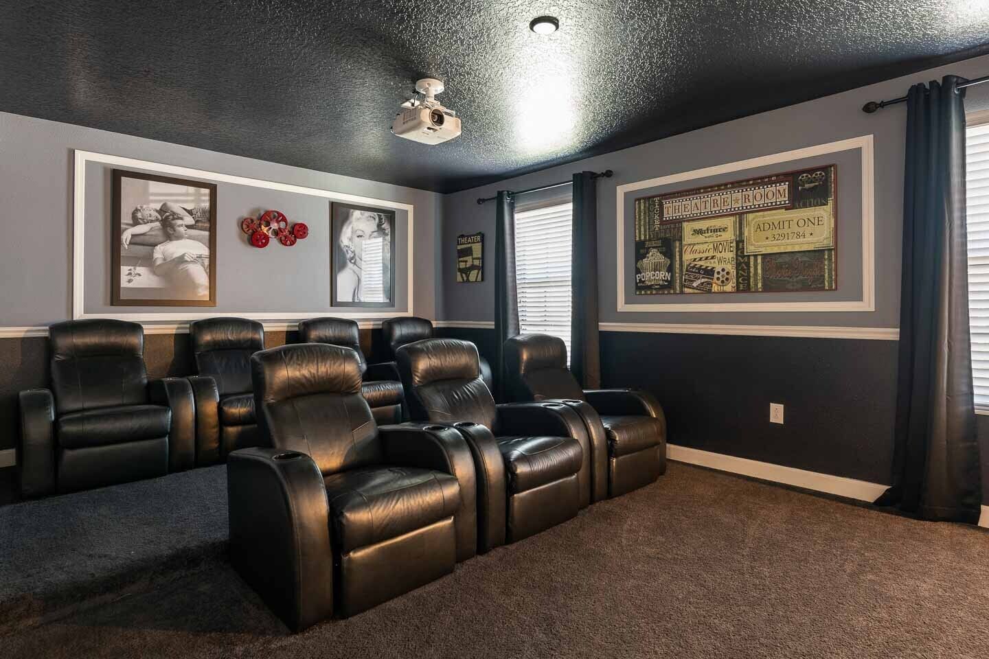 Una sala de cine en casa con sillones de cuero reclinables, un proyector de techo, carteles de películas enmarcados y una ventana con persianas.