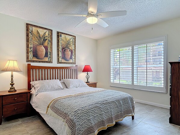 A bedroom with a bed, two nightstands, a ceiling fan, a window with blinds, and two framed paintings on the wall.