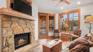 Un salon chaleureux avec une cheminée en pierre, des meubles en bois et de grandes fenêtres donnant sur l’extérieur.
