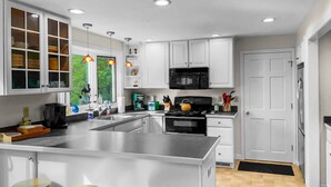 A modern kitchen with white cabinets, a large island with stools, a black microwave, and a view of greenery outside. - The Heron's Nest - Sea Breezes & Sweet Dreams (Chatham)