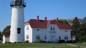 A U.S. Coast Guard Station Chatcum building with a white exterior and red roof, adjacent to a white lighthouse. - The Heron's Nest - Sea Breezes & Sweet Dreams (Chatham)