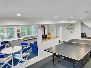 A modern living room with a ping pong table, a blue sofa, and a window with a view of greenery.