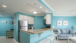 Une cuisine avec des meubles blancs, un réfrigérateur en acier inoxydable et un plan de travail en granit. Un mur bleu est décoré d’une enseigne « PLAGE » et de deux tableaux encadrés.