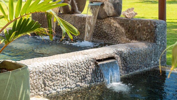 A stone water feature with a small waterfall, surrounded by greenery and a view of the ocean. - Aloha Aku Ohana Suite – Spacious Garden View Oasis (Kihei)