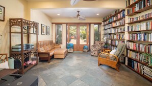 A spacious living room with a leather sofa, a bookshelf filled with books, a ceiling fan, and a view through the glass door to the outside. - Aloha Aku Ohana Suite – Spacious Garden View Oasis (Kihei)