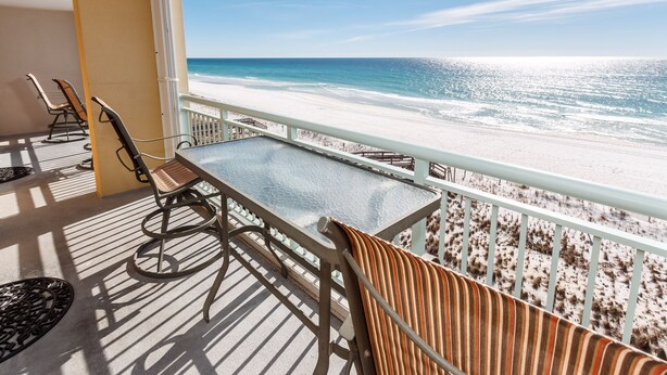 A balcony with a glass table and chairs overlooking a beach.