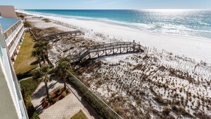 A beach view from a hotel balcony with a wooden walkway leading to the beach, palm trees, and a clear sky.