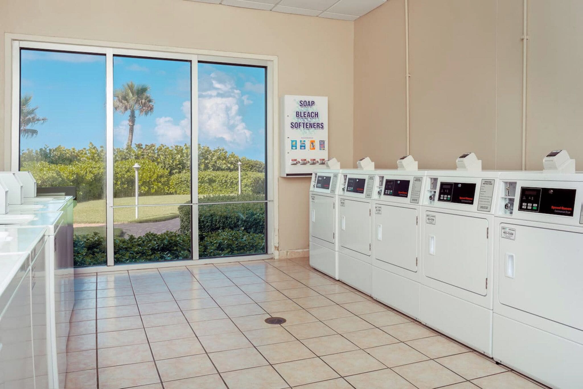 A laundry facility with multiple washing machines, a sign listing detergent types, and a view of greenery through large windows.