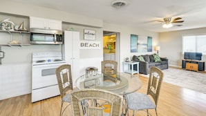 A modern kitchen with white cabinets, a microwave, and a stove. There is a dining area with wicker chairs and a glass table. A living room with a gray sofa and a TV is visible. - Walk to Gulf: Surfside Beach Home w/ Deck (Surfside Beach)