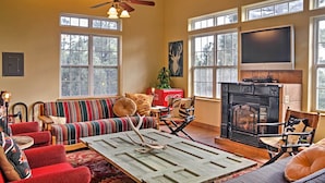A cozy living room with a fireplace, a large window, and a ceiling fan. - Quiet Ruidoso Hideaway - 2 Mi to Grindstone Lake! (Ruidoso)