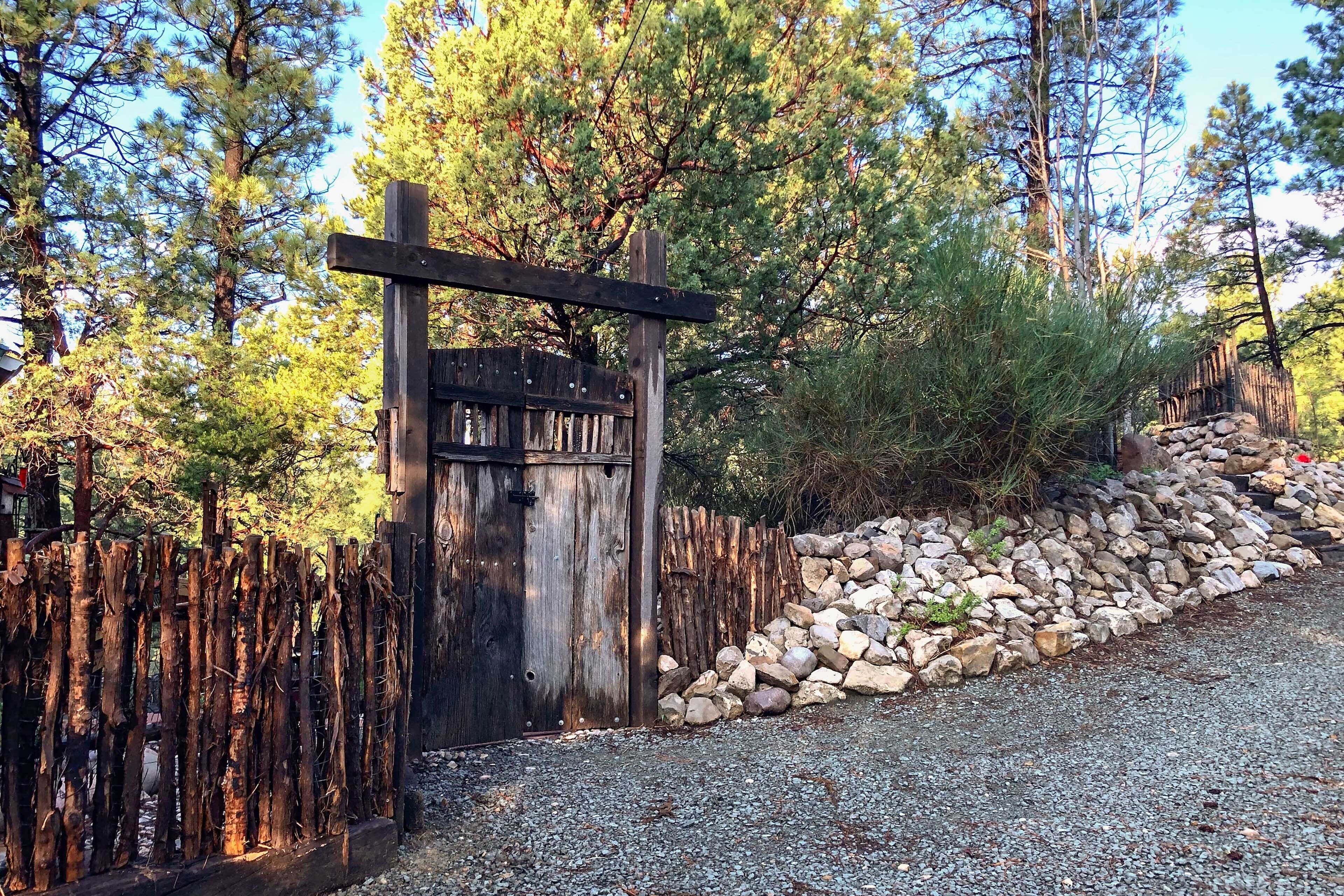 A rustic wooden gate with a sign, surrounded by a wooden fence and a stone wall, leading into a wooded area.