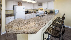 A kitchen with white cabinets, granite countertops, a refrigerator, stove, microwave, and bar stools. - IBR 3006: Sandy Smiles - Unbeatable Location, Unmatched Oceanfront Views! (Fort Walton Beach)