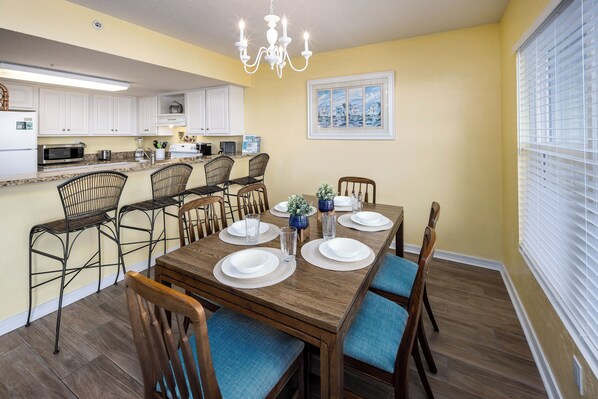 A dining area with a wooden table set for six, surrounded by wicker chairs with blue cushions.