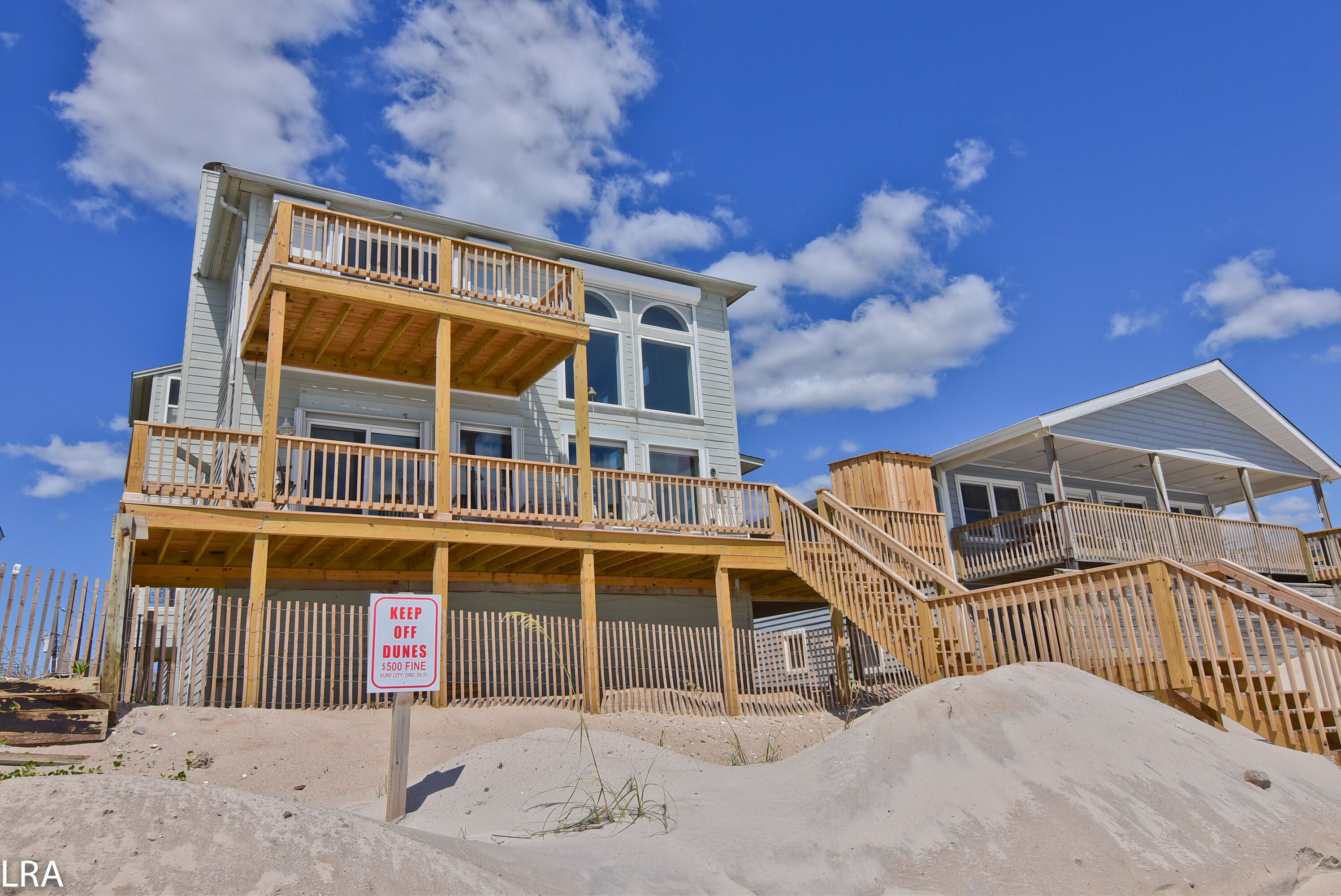 Une maison de plage à deux étages, dotée d’une terrasse en bois et d’un panneau indiquant « Défense de marcher sur les dunes ».