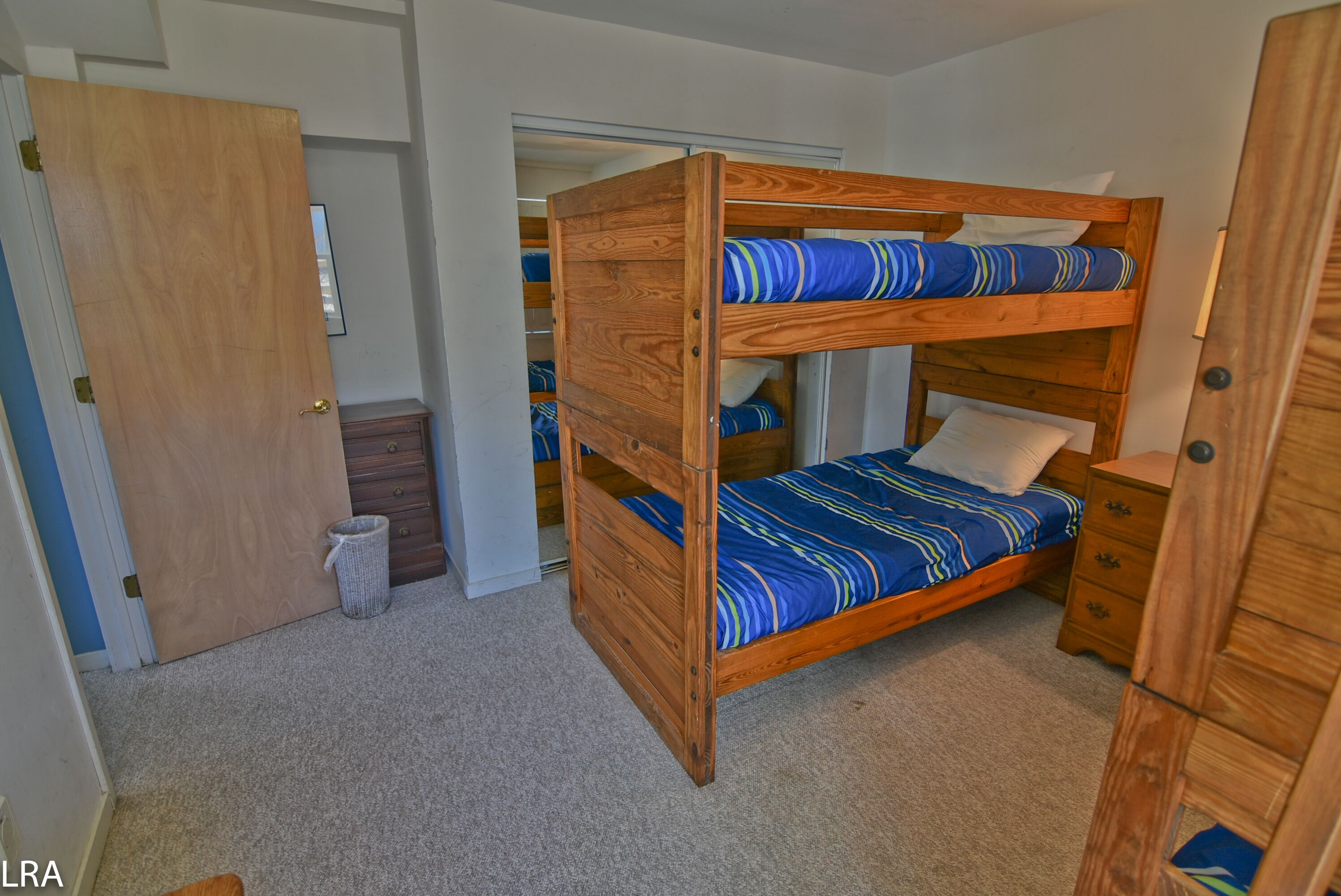 A bunk bed room with a wooden bunk bed, a small wooden nightstand, a dresser, and a door leading to another room.