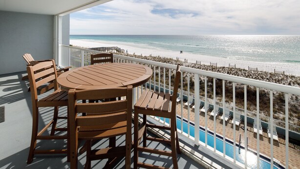 Ein Balkon mit Holztisch und Stühlen, der Blick auf einen Strand und ein Schwimmbad freigibt.