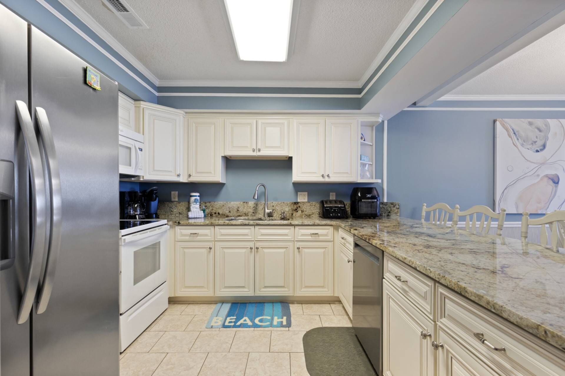 Una cocina moderna con armarios blancos, un refrigerador de acero inoxidable, una encimera de granito y una alfombra azul con la palabra 'BEACH'.