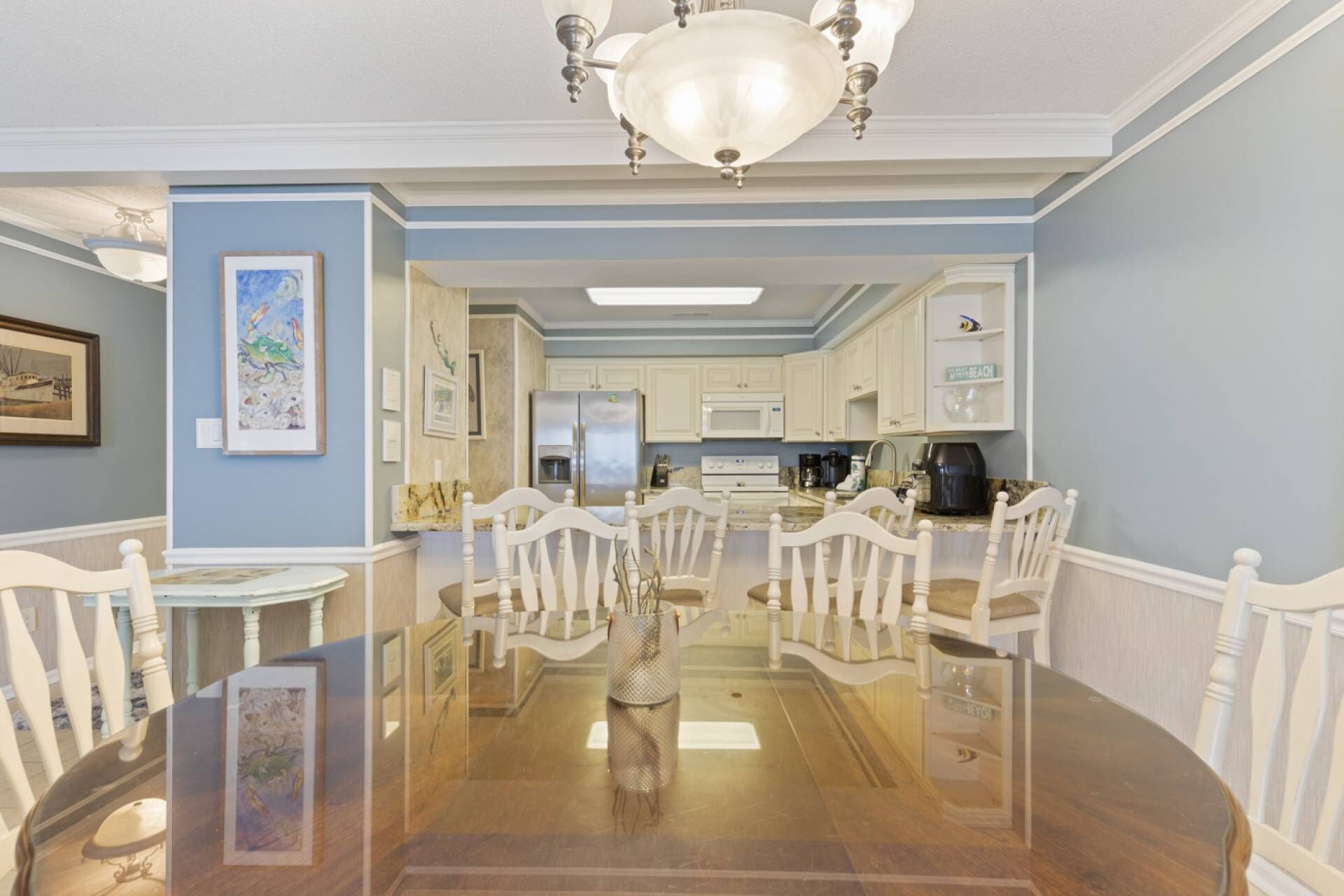A dining area with a glass table and white chairs, a kitchen with white cabinets, and a chandelier.