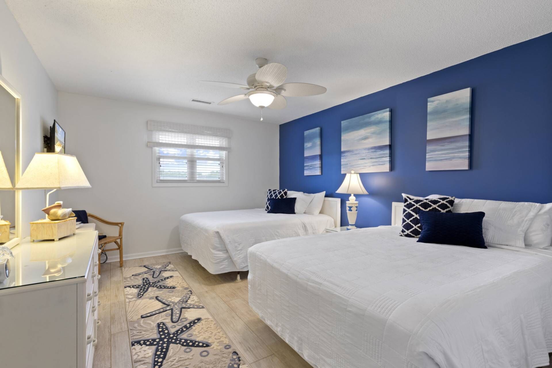 Une chambre avec deux lits, un ventilateur de plafond, une fenêtre avec des stores et un tapis orné de motifs d’étoiles de mer.