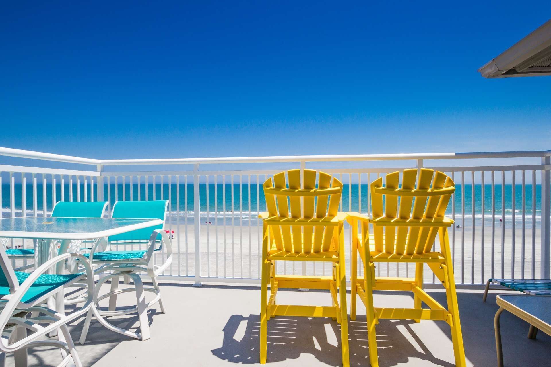 A balcony with a view of the ocean, featuring two yellow Adirondack chairs and a table with blue cushions.