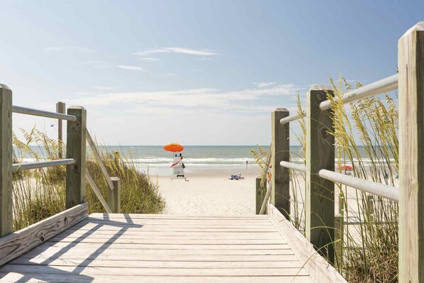 A wooden boardwalk leading to a beach with a person under an umbrella.