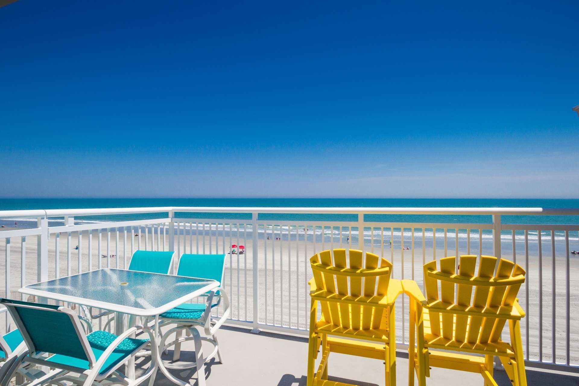 Ein Balkon mit Glastisch und Stühlen, der Blick auf einen Strand mit blauem Himmel bietet.