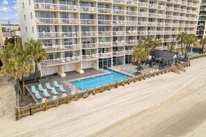 A multi-story hotel with balconies, a pool area with lounge chairs, and a sandy beach.