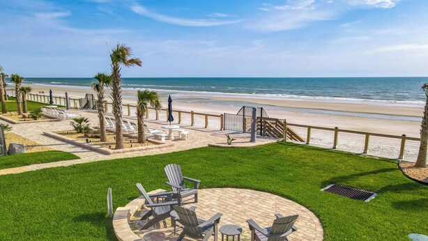 A patio area with Adirondack chairs and a fire pit, overlooking a beach with palm trees and a fence.