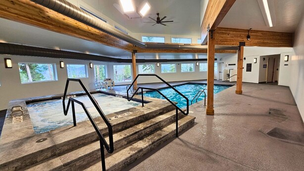 An indoor swimming pool with steps leading into it, wooden beams on the ceiling, and windows with a view of trees.